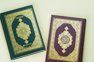 Two ornate books with intricate gold patterns on the covers; one book is green and the other is maroon. The green book has an elaborate symmetrical design with Arabic script, while the maroon book features a similar decorative style. Both cover designs include floral motifs and geometric shapes.