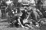 A group of emergency responders, including firefighters and paramedics wearing protective gear and masks, are attending to an individual lying on the ground. The scene takes place outdoors by a fence with emergency vehicles in the background.
