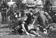A group of emergency responders, including firefighters and paramedics wearing protective gear and masks, are attending to an individual lying on the ground. The scene takes place outdoors by a fence with emergency vehicles in the background.