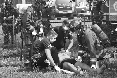 A group of emergency responders, including firefighters and paramedics wearing protective gear and masks, are attending to an individual lying on the ground. The scene takes place outdoors by a fence with emergency vehicles in the background.