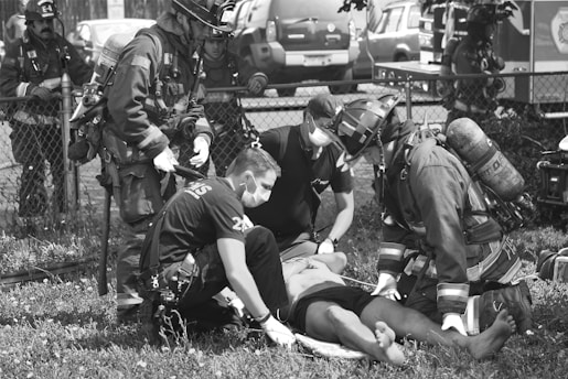 A group of emergency responders, including firefighters and paramedics wearing protective gear and masks, are attending to an individual lying on the ground. The scene takes place outdoors by a fence with emergency vehicles in the background.