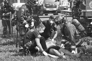 A group of emergency responders, including firefighters and paramedics wearing protective gear and masks, are attending to an individual lying on the ground. The scene takes place outdoors by a fence with emergency vehicles in the background.