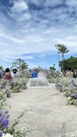 A beautifully arranged outdoor wedding ceremony with a couple standing at the altar surrounded by flowers. Guests are seated along a path leading to the couple. The sky is partly cloudy, adding a serene backdrop to the scene with palm trees visible in the distance.
