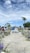 A beautifully arranged outdoor wedding ceremony with a couple standing at the altar surrounded by flowers. Guests are seated along a path leading to the couple. The sky is partly cloudy, adding a serene backdrop to the scene with palm trees visible in the distance.