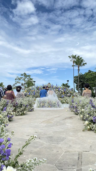 A sunlit outdoor wedding ceremony with elegant floral arrangements and guests seated under delicate white canopies.