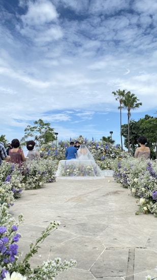 A beautifully arranged outdoor wedding ceremony with a couple standing at the altar surrounded by flowers. Guests are seated along a path leading to the couple. The sky is partly cloudy, adding a serene backdrop to the scene with palm trees visible in the distance.