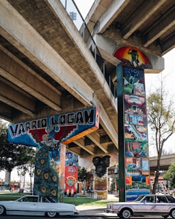 Colorful murals adorn the concrete pillars of an urban overpass. The artwork displays vibrant and detailed images depicting cultural and community themes, featuring elements such as peacocks and figures engaged in various activities. Below the overpass, classic cars are parked on the side of the road, and trees and grass add a touch of greenery.