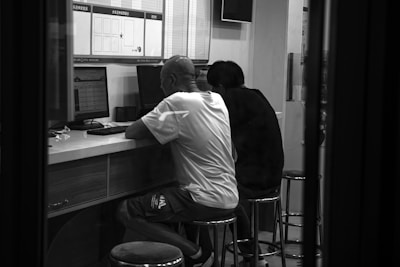 Two people are seated on stools at a counter, focused on computer screens in what appears to be an internet cafe or office setting. The room is minimally decorated, and both individuals are facing away from the camera, engrossed in their tasks.