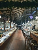 A spacious indoor market with high, arched ceilings and an array of goods displayed on either side of a central aisle. Shelves on the left are lined with various packaged products, while the right showcases a deli counter with fresh items. Wooden stools are arranged along the counter area. Bright lights illuminate the scene, and a large, visible clock hangs from the ceiling.