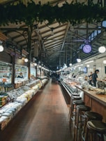 A spacious indoor market with high, arched ceilings and an array of goods displayed on either side of a central aisle. Shelves on the left are lined with various packaged products, while the right showcases a deli counter with fresh items. Wooden stools are arranged along the counter area. Bright lights illuminate the scene, and a large, visible clock hangs from the ceiling.