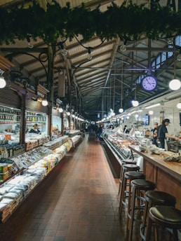 A spacious indoor market with high, arched ceilings and an array of goods displayed on either side of a central aisle. Shelves on the left are lined with various packaged products, while the right showcases a deli counter with fresh items. Wooden stools are arranged along the counter area. Bright lights illuminate the scene, and a large, visible clock hangs from the ceiling.