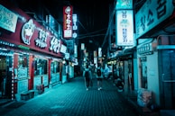 A nighttime street scene in a bustling area with neon signs in Korean hanging from buildings. The lighting is predominantly blue, casting a moody glow over the narrow street. People casually walk along the brick-paved road, passing by vibrant storefronts on either side.
