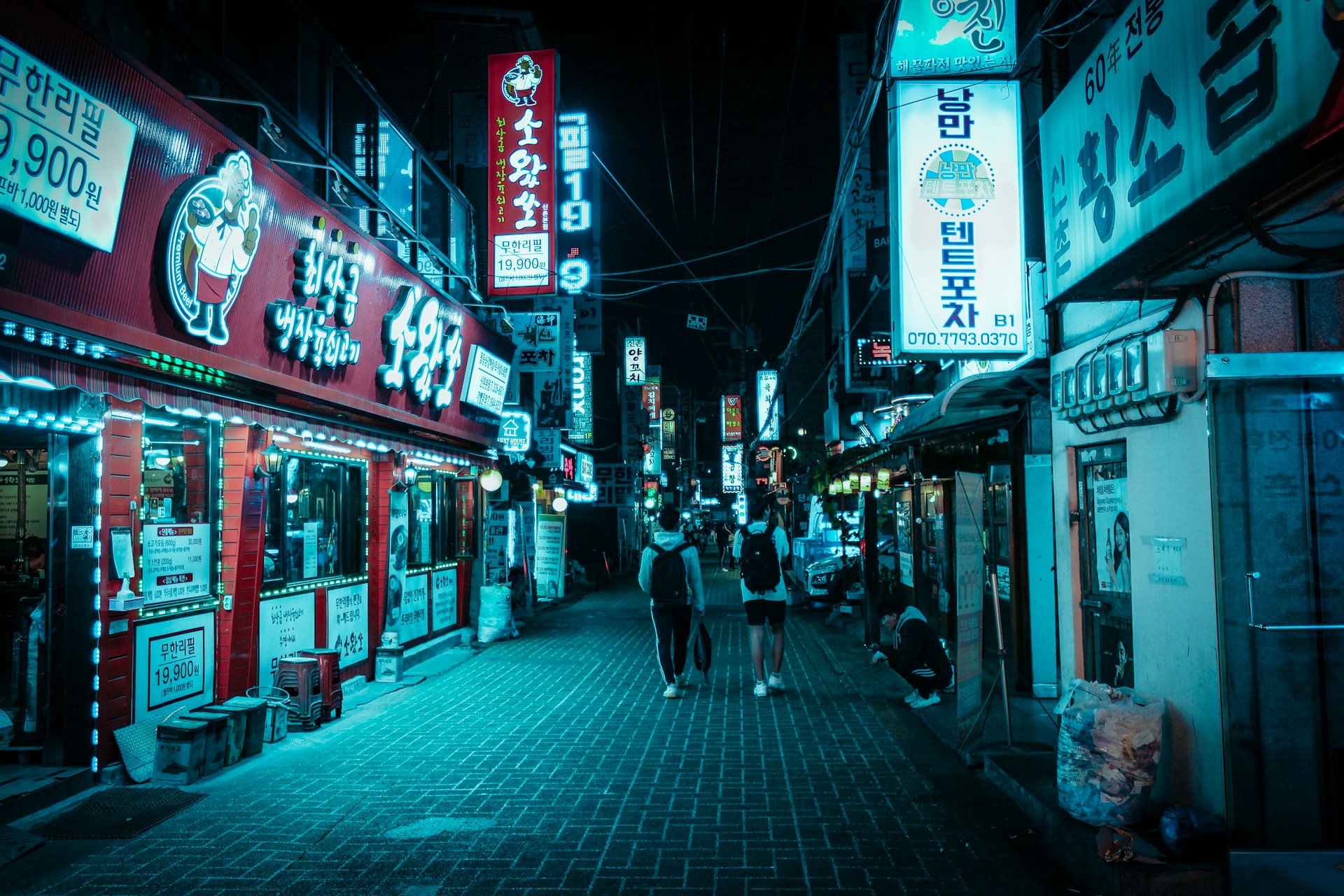 A nighttime street scene in a bustling area with neon signs in Korean hanging from buildings. The lighting is predominantly blue, casting a moody glow over the narrow street. People casually walk along the brick-paved road, passing by vibrant storefronts on either side.