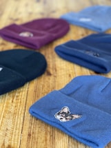 Several knitted beanies are laid out on a wooden surface. Each beanie has a distinct color and features an embroidered patch of an animal on the front. The focus is on the beanie in the foreground, which is blue with a small dog patch. Other colors visible include black, maroon, and light blue.