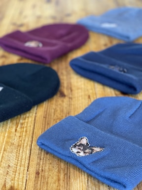 Several knitted beanies are laid out on a wooden surface. Each beanie has a distinct color and features an embroidered patch of an animal on the front. The focus is on the beanie in the foreground, which is blue with a small dog patch. Other colors visible include black, maroon, and light blue.