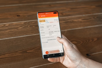 A hand holding a smartphone displaying a financial trading app interface with details of a sell order for MSFT Nasdaq, including current bid, ask prices, quantity, and order type. The background is a wooden surface with a visible grain pattern.