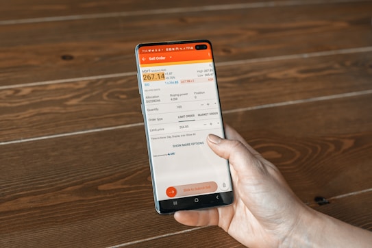 A hand holding a smartphone displaying a financial trading app interface with details of a sell order for MSFT Nasdaq, including current bid, ask prices, quantity, and order type. The background is a wooden surface with a visible grain pattern.