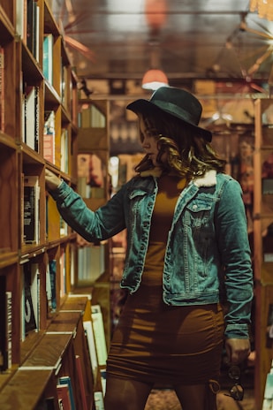 A woman stands in a cozy, dimly lit bookstore aisle, wearing a denim jacket and a wide-brimmed hat. She is reaching out towards a bookshelf filled with colorful books, intently examining them. The atmosphere is warm, with reflections of ambient lights above.