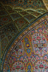 An intricate and vibrant mosaic pattern covers the surface, featuring a mix of geometric and floral designs. The artwork includes an array of colors and detailed craftsmanship, with floral motifs and Arabic script embedded within the pattern.