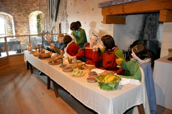 A group of mannequins dressed in medieval attire is seated around a long table, which is laden with various foods and drinks, including bread, fruit, and meats. They are positioned in an animated discussion environment in a rustic room with stone walls, a wooden floor, and an open fireplace.