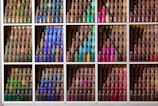 A collection of small bottles filled with colorful materials such as sand or spices in various shades. These bottles are neatly arranged in a grid of compartments creating a visually striking display. The colors range from greens and yellows to blues, purples, pinks, and reds, each compartment containing a specific color scheme.