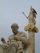 A detailed marble statue of an ancient philosopher seated, juxtaposed with a towering statue of a goddess holding a spear and shield on a column with ornate gold accents.