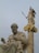A detailed marble statue of an ancient philosopher seated, juxtaposed with a towering statue of a goddess holding a spear and shield on a column with ornate gold accents.