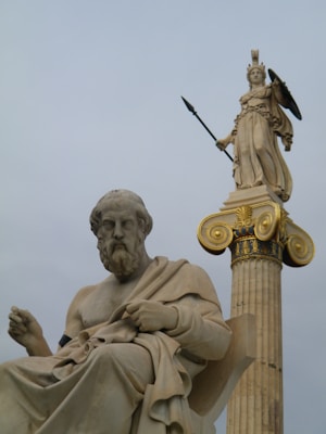A detailed marble statue of an ancient philosopher seated, juxtaposed with a towering statue of a goddess holding a spear and shield on a column with ornate gold accents.