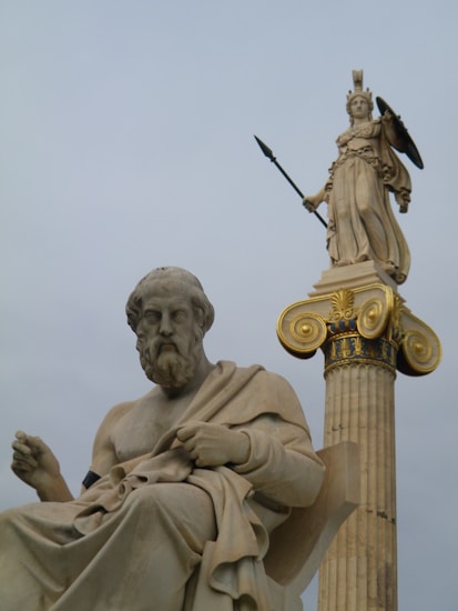 A detailed marble statue of an ancient philosopher seated, juxtaposed with a towering statue of a goddess holding a spear and shield on a column with ornate gold accents.