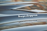 Glossy black car surface with a polished silver emblem displaying the word 'Superleggera'. The image captures the smooth curves and fine details of the car's design.