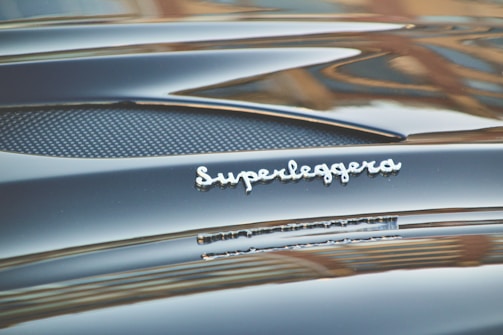 Glossy black car surface with a polished silver emblem displaying the word 'Superleggera'. The image captures the smooth curves and fine details of the car's design.