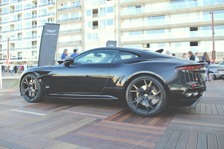 A sleek black sports car is parked on a wooden platform in an urban area. The car's design is modern and aerodynamic, with large alloy wheels and a shiny exterior. People are gathered around in the background, along with a tall banner featuring a logo. The backdrop includes a multi-story building with numerous windows.