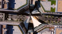 An aerial view of a modern architectural complex featuring a symmetrical layout with angular rooftops. The central area has a large white canopy covering a pedestrian walkway lined with trees and greenery. The surrounding buildings have flat rooftops, some with patches of greenery or tiles.