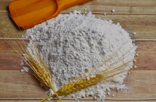 A pile of white flour sits on a wooden surface, accompanied by a wooden scoop and two sprigs of wheat.