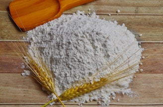 A pile of white flour sits on a wooden surface, accompanied by a wooden scoop and two sprigs of wheat.