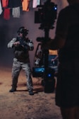 A person wearing tactical gear and night-vision goggles holds a firearm while standing in a dimly-lit environment. The background includes a vehicle with its door open and clothes hanging overhead. A camera rig is partially visible, suggesting a film or photo shoot setting.