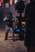 A person wearing tactical gear and night-vision goggles holds a firearm while standing in a dimly-lit environment. The background includes a vehicle with its door open and clothes hanging overhead. A camera rig is partially visible, suggesting a film or photo shoot setting.