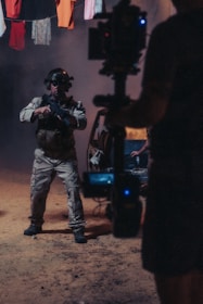 A person wearing tactical gear and night-vision goggles holds a firearm while standing in a dimly-lit environment. The background includes a vehicle with its door open and clothes hanging overhead. A camera rig is partially visible, suggesting a film or photo shoot setting.