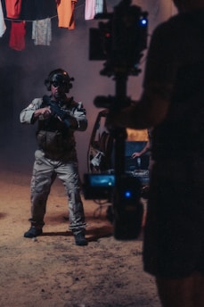 A person wearing tactical gear and night-vision goggles holds a firearm while standing in a dimly-lit environment. The background includes a vehicle with its door open and clothes hanging overhead. A camera rig is partially visible, suggesting a film or photo shoot setting.