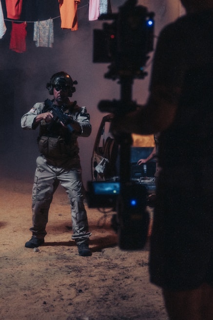 A person wearing tactical gear and night-vision goggles holds a firearm while standing in a dimly-lit environment. The background includes a vehicle with its door open and clothes hanging overhead. A camera rig is partially visible, suggesting a film or photo shoot setting.