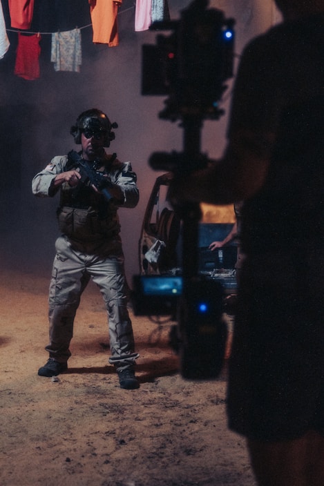 A person wearing tactical gear and night-vision goggles holds a firearm while standing in a dimly-lit environment. The background includes a vehicle with its door open and clothes hanging overhead. A camera rig is partially visible, suggesting a film or photo shoot setting.