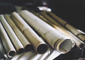 Several rolled-up sheets of paper are placed on a surface, likely containing architectural plans or blueprints. The image is focused on the cylindrical form of the scrolls, showing the textured and slightly yellowed paper. The background is dimly lit, adding a sense of age or antiquity to the scene.
