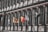An architectural facade features a series of tall, dark columns and extensive windows. In front of the building, several flags are flying, including a black and yellow flag with a lion emblem, a red and yellow flag, and others in black and white designs.