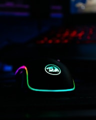 A gaming mouse with RGB lighting, featuring vibrant colors including purple, green, and blue. The mouse has a logo on it and is illuminated against a dark background, with a keyboard in the distance emitting a red glow.