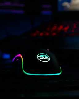 A gaming mouse with RGB lighting, featuring vibrant colors including purple, green, and blue. The mouse has a logo on it and is illuminated against a dark background, with a keyboard in the distance emitting a red glow.
