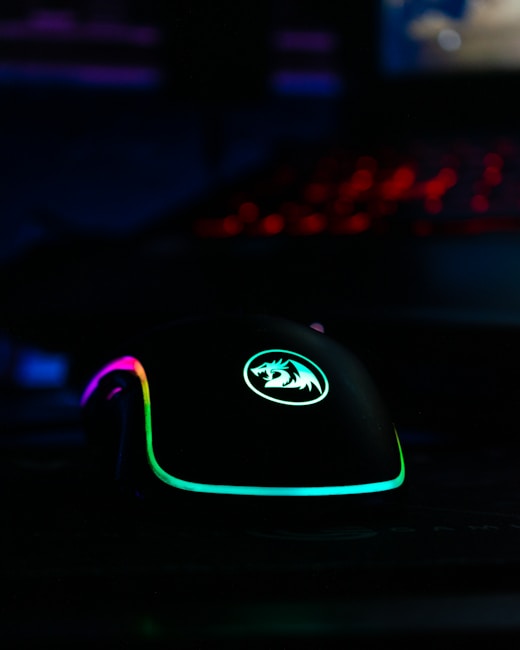 A gaming mouse with RGB lighting, featuring vibrant colors including purple, green, and blue. The mouse has a logo on it and is illuminated against a dark background, with a keyboard in the distance emitting a red glow.