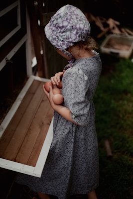 A child wearing a floral-patterned bonnet and dress is holding several eggs, standing by a wooden chicken coop. The scene suggests a rural or farm setting, with grass visible in the foreground.