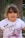 A young girl with long hair is wearing a white t-shirt with the text 'FLY HIGH' and a graphic of a winged roller skate in pink. She appears to be outdoors with a tree trunk and some metal bars in the background.