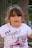 A young girl with long hair is wearing a white t-shirt with the text 'FLY HIGH' and a graphic of a winged roller skate in pink. She appears to be outdoors with a tree trunk and some metal bars in the background.
