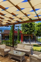 A cozy outdoor seating area featuring wicker furniture arranged under a wooden pergola with a crisscross pattern of beige canvas providing shade. The scene is set against a lush background of trees and shrubs, and a building with a modern facade is visible. Sunlight casts shadows across the furniture and the wooden floor.